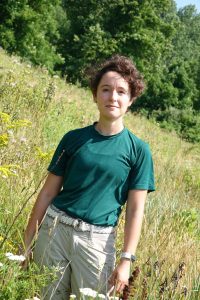 Jordan Coscia standing in field of tall grasses with tree line in the background. Jordan is wearing tan slacks and a green t-shirt. 